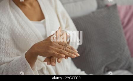 Jeune femme afro-américaine se frottant les mains dans un salon confortable portant une robe blanche à la maison. Banque D'Images