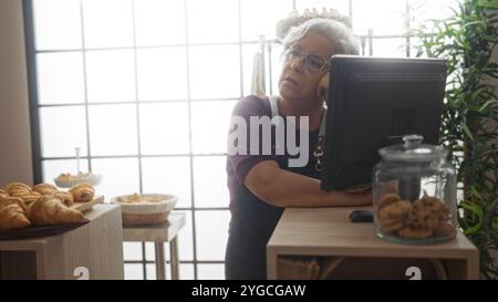 Femme mature avec les cheveux gris debout derrière un comptoir dans une boulangerie avec des pâtisseries et des biscuits, l'air réfléchi Banque D'Images