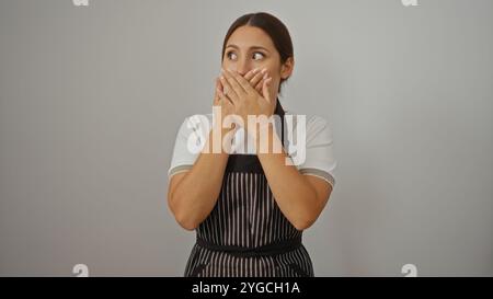 Jeune belle femme brune portant tablier sur fond blanc regardant surpris avec les mains couvrant la bouche Banque D'Images