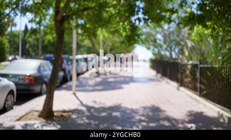 Vue défocalisée d'un chemin extérieur bordé de voitures garées et ombragé par des arbres avec des gens flous en arrière-plan Banque D'Images