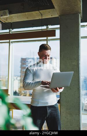 Un jeune homme dans un espace de travail moderne se tient près d'une grande fenêtre. Un homme en vêtements décontractés avec un ordinateur portable dans les mains. Télétravail. Gentil jeune manager Banque D'Images