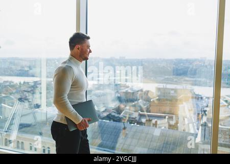 Un jeune homme dans un espace de travail moderne se tient près d'une grande fenêtre. Un homme en vêtements décontractés avec un ordinateur portable dans les mains. Télétravail. Gentil jeune manager Banque D'Images