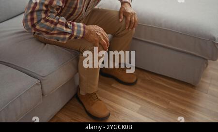 Un homme âgé est assis sur un canapé dans un salon confortable, les mains reposant sur ses genoux, mettant en valeur un environnement familial détendu. Banque D'Images