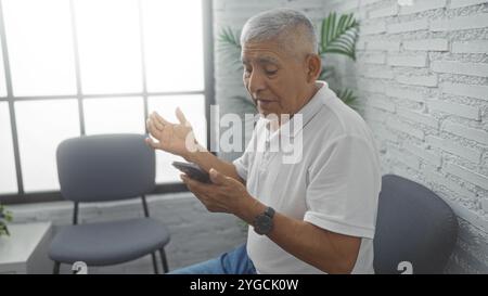 Un homme hispanique d'âge moyen en chemise blanche tient un téléphone assis dans une salle d'attente intérieure lumineuse avec des chaises grises et des murs de briques blanches. Banque D'Images