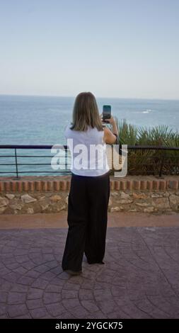 Jeune femme en haut blanc et pantalon noir prenant une photo de l'océan avec son smartphone à l'extérieur près d'une balustrade en pierre et d'une verdure luxuriante sur un da ensoleillé Banque D'Images