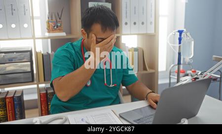 Jeune médecin chinois dans une chambre d'hôpital, paraissant stressé, portant des gommages avec un stéthoscope, assis à un bureau avec un ordinateur portable, dossier médical, et Banque D'Images