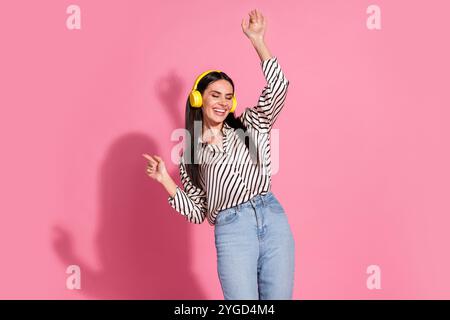 Jeune femme en chemise rayée appréciant la musique avec des écouteurs jaunes sur fond rose Banque D'Images