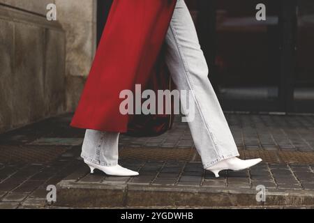 Gros plan sur les jambes d'une femme élégante et méconnaissable marchant dans la rue urbaine, portant un Jean, un manteau Bordeaux et des bottes à talons blancs à la mode, capturant cit Banque D'Images