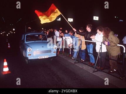ARCHIVE PHOTO : il y a 35 ans, le 9 novembre 1989, le mur de Berlin est tombé. Un chauffeur Trabant d'Allemagne de l'est traverse un poste frontalier dans l'après-midi (du 9 au 10 novembre 1989). Sur le côté droit de la route, derrière une barrière, les gens applaudissent et font signe. Un homme agite un drapeau noir-rouge-or. Format paysage. ? Banque D'Images