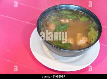 Soupe traditionnelle thaïlandaise épicée Tom yum avec poulet servie dans un bol noir et sur un plat blanc avec une table en bois rose Banque D'Images