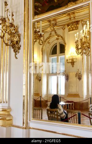 Vienne, Autriche - 6 octobre 2024 : intérieur doré du palais avec miroir reflétant une visiteuse assise, avec architecture baroque, lustres, Banque D'Images