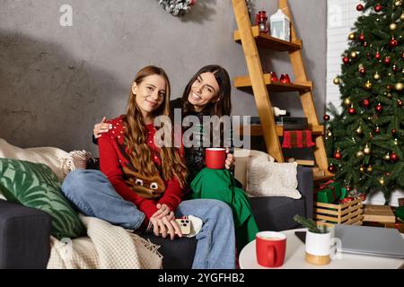 Deux copines apprécient la compagnie de l'autre dans des chandails confortables tout en célébrant la saison des fêtes à la maison. Banque D'Images