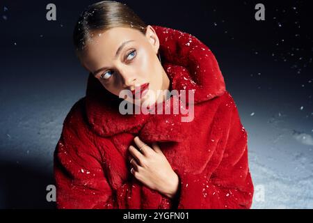 Une jeune femme élégante pose gracieusement dans un manteau rouge vif tandis que la neige tombe doucement autour d'elle. Banque D'Images