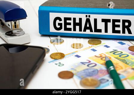 Bavière, Allemagne - 7 novembre 2024 : les salaires sont écrits sur un classeur bleu à côté des billets et des pièces en euros sur un bureau. Image symbolique de l'augmentation des salaires et des paiements salariaux en Allemagne. PHOTOMONTAGE *** Gehälter steht auf einem blauen Aktenordner neben Geld-Scheinen und Euro-Münzen auf einem Schreibtisch. Symbolbild zu Gehalt und Gehaltszahlungen Steigend in Deutschland. FOTOMONTAGE Banque D'Images