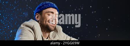 Un jeune homme souriant embrasse la joie hivernale avec des flocons de neige tourbillonnant autour de lui à l'intérieur. Banque D'Images