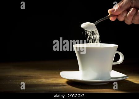 Main masculine mettant de nombreuses cuillères à café pleines de sucre granulé dans la tasse blanche et remuant une boisson, gros plan. Risque sanitaire et concept de diabète Banque D'Images