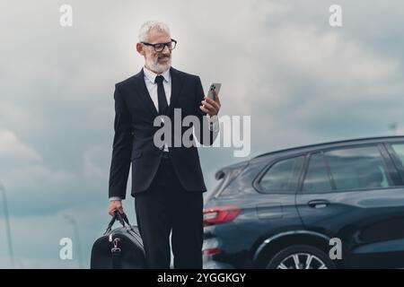 Homme d'affaires senior en costume formel à l'extérieur sur la rue de la ville près de la voiture avec smartphone et sac de voyage Banque D'Images