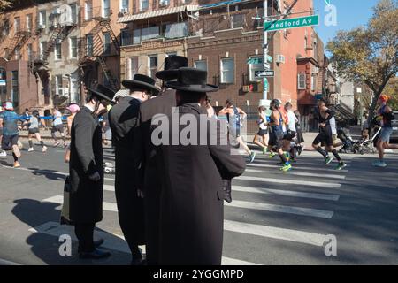 Aux 10 miles du marathon de New York de 2024, les hommes juifs orthodoxes attendent l'occasion de traverser Bedford Ave à l'amiudst de l'immense couronne de coureurs. Banque D'Images