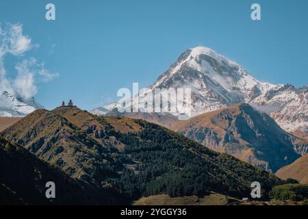 Gergeti Trinity Church perchée sur une colline avec le mont Kazbek enneigé dominant en arrière-plan, sous un ciel bleu vibrant en Géorgie Banque D'Images