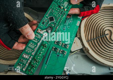 Réparateur portant des gants inspectant une carte de circuit imprimé cassée d'une cuisinière électrique pendant un travail d'entretien ou de réparation Banque D'Images