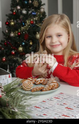 Adorable petite fille profitant des vacances d'hiver à la maison, portant un pull de Noël, des biscuits en pain d'épices sur la table, ambiance festive chaleureuse. Banque D'Images