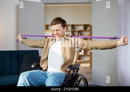 Jeune homme handicapé en fauteuil roulant faisant des exercices avec un élastique à la maison. Un homme handicapé de race blanche s'entraîner dans la salle de séjour. Banque D'Images