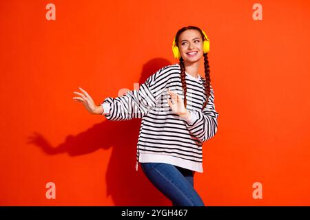 Jeune femme aux cheveux rouges portant des écouteurs et un pull rayé danse joyeusement sur un fond orange. Banque D'Images