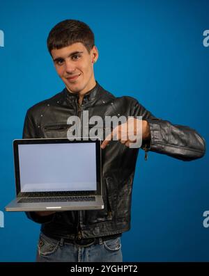 Jeune programmeur pointant vers son ordinateur portable avec écran blanc, isolé sur fond bleu de studio Banque D'Images