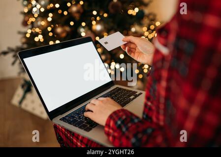Shopping de vacances en ligne avec un ordinateur portable et une carte de crédit, lumières de sapin de Noël confortables en arrière-plan, ambiance festive. Banque D'Images