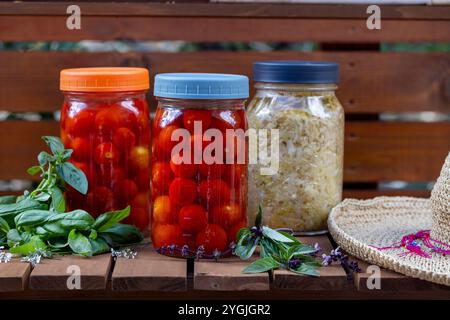 Comptoir en bois de chariot de jardin et bocaux maçon avec tomates cerises en fermentation et chou, entouré de basilic frais et de feuilles de basilic thaï et chapeau de paille Banque D'Images