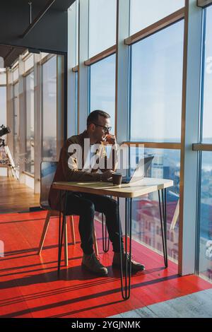 Un homme équilibré dans des lunettes et des vêtements décontractés travaille sur un ordinateur portable à partir d'un espace de travail confortable. Un freelance performant travaille à distance. Banque D'Images