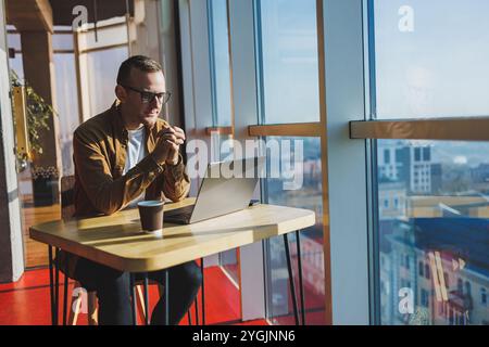 Un homme équilibré dans des lunettes et des vêtements décontractés travaille sur un ordinateur portable à partir d'un espace de travail confortable. Un freelance performant travaille à distance. Banque D'Images