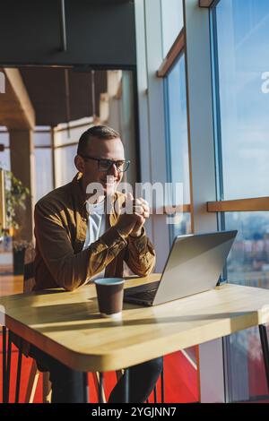 Un homme équilibré dans des lunettes et des vêtements décontractés travaille sur un ordinateur portable à partir d'un espace de travail confortable. Un freelance performant travaille à distance. Banque D'Images