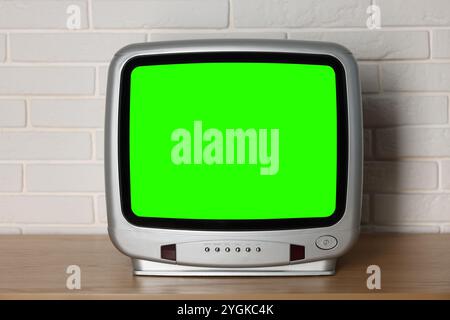 Téléviseur rétro avec écran vert sur table en bois près du mur de briques blanches. Touche Chroma Banque D'Images