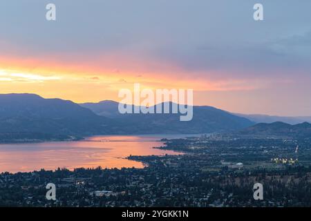 Une vue imprenable sur Kelowna et le lac Okanagan au coucher du soleil, avec des couleurs chaudes illuminant le ciel et se reflétant sur l'eau tranquille. Banque D'Images