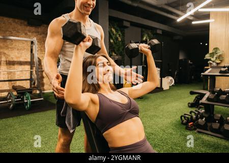 Une femme soulevant des haltères avec le soutien de son entraîneur dans un gymnase, incarnant la force, la motivation et les objectifs de fitness. Banque D'Images