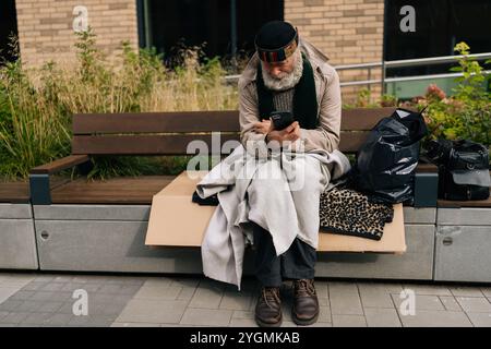 Portrait d'un homme adulte mature assis sur le banc de la ville, en tapant smartphone au milieu de la solitude urbaine, éclairant sur la pauvreté et l'isolement dans le vieillissement Banque D'Images