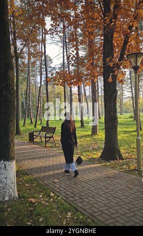 Belle jeune femme portant un manteau noir marche le long de l'allée des arbres colorés dans le parc d'automne. Rayons de soleil chauds allumés l'orange laisse un banc et la Banque D'Images