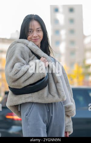 Une jeune belle femme de 20-25 ans avec les cheveux longs marche le long d'une rue de la ville dans un manteau de fourrure confortable et un pantalon gris lâche, avec un sac à main en h. Banque D'Images