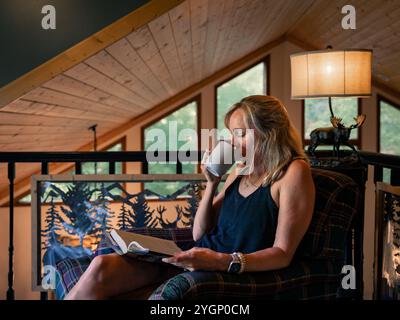 Une femme est assise confortablement dans une cabine confortable, sirotant une boisson chaude tout en lisant son livre par les fenêtres. Banque D'Images