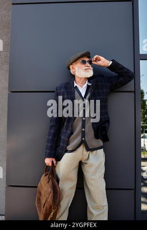 Un homme senior à la mode pose en toute confiance dans un blazer à carreaux et une casquette plate près d'un bâtiment contemporain. Banque D'Images