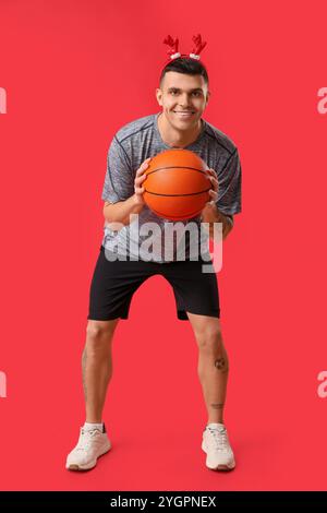 Jeune homme heureux dans des cornes de renne jouant au basket-ball sur fond rouge Banque D'Images