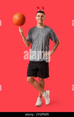 Jeune homme heureux dans des cornes de renne avec basket-ball sur fond rouge Banque D'Images