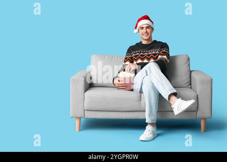 Jeune homme dans le chapeau de Santa avec seau de pop-corn regardant le film et assis sur le canapé sur fond bleu. Fête de Noël Banque D'Images