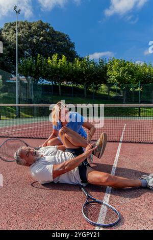 la joueuse de tennis senior verticale s'agenouille à côté de son partenaire blessé sur le court, offrant confort et soutien alors qu'il tient sa jambe dans la douleur Banque D'Images