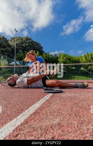 le joueur de tennis âgé vertical est allongé sur le court, serrant sa jambe et grimacant de douleur, tandis que son partenaire s'agenouille à côté de lui pour offrir du confort Banque D'Images