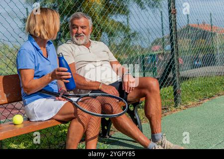 gros plan souriant joueur de tennis senior engage une conversation amicale avec son partenaire tout en étant assis sur un banc pendant une pause match Banque D'Images