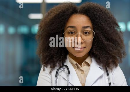 Médecin afro-américain confiant en manteau blanc avec stéthoscope, cheveux bouclés et lunettes dans un cadre médical moderne. Fait preuve de professionnalisme, d'expertise et de compassion. Banque D'Images