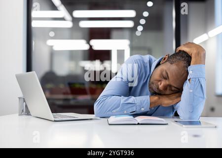 Homme d'affaires afro-américain mature dormant au bureau dans un bureau moderne. Professionnel fatigué portant chemise bleue reposant avec ordinateur portable, livre ouvert, téléphone sur la table. Surmenage, fatigue, concept de stress. Banque D'Images