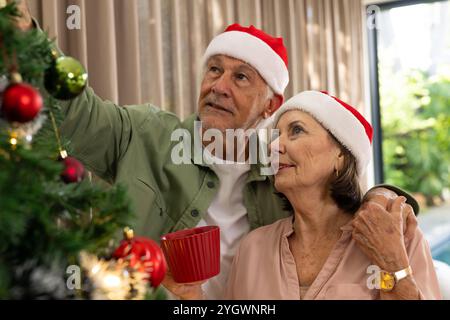 Couple senior décorant l'arbre de Noël à la maison, portant des chapeaux de Père Noël, souriant chaleureusement Banque D'Images